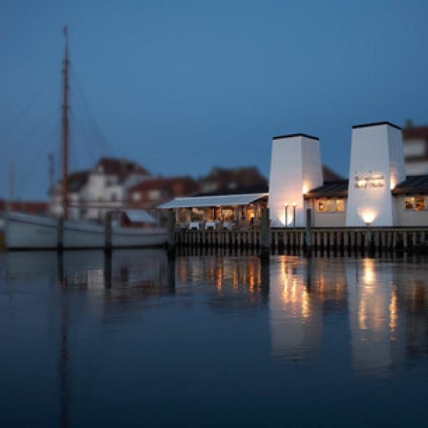 Hvid facade ud til en havn, hvor der ligger skibe foran, og det er aften med tændte lys