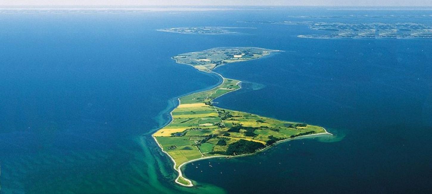 Dronefoto af Avernakø i skarpe, grønne farver med dybblåt vand hele vejen rundt om og horisonten i baggrunden. 