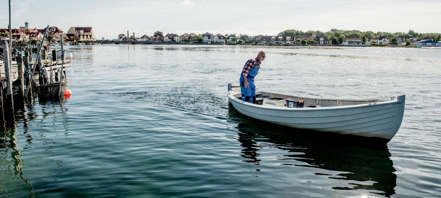 Hvid båd med mand i ternet skjorte ombord sejler ud fra Kerteminde Havn på roligt vand. Byen ses i baggrunden i horisonten.