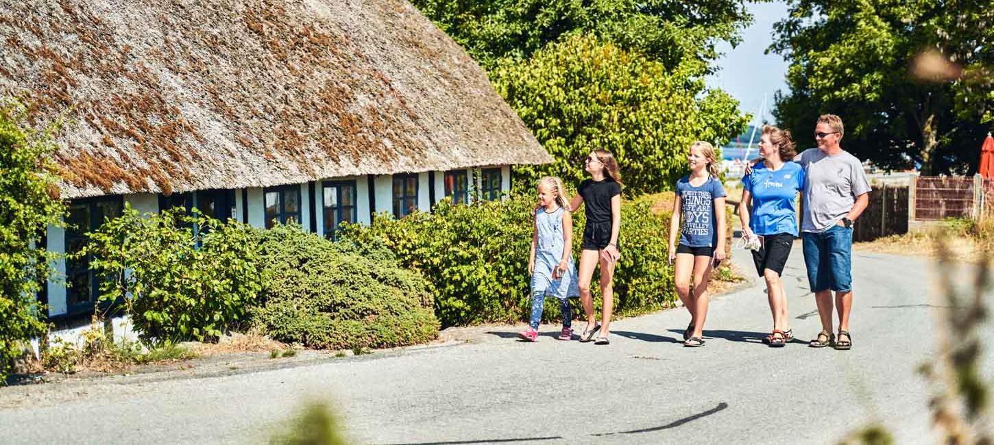 I fuld solskin går en familie med tre børn på række. De går på en asfaltvej. I baggrunden omfavner træer vejen og i venstre side ses et hvidt bindingsværks hus med stråtag. 