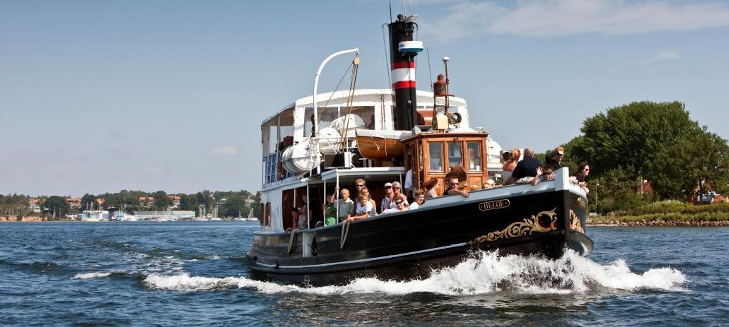 En gammel båd sejler på havet. I stævnen af båden står en masse turister og spejder udover havet. I baggrunden kan man se vandkanten fyldt med træer.