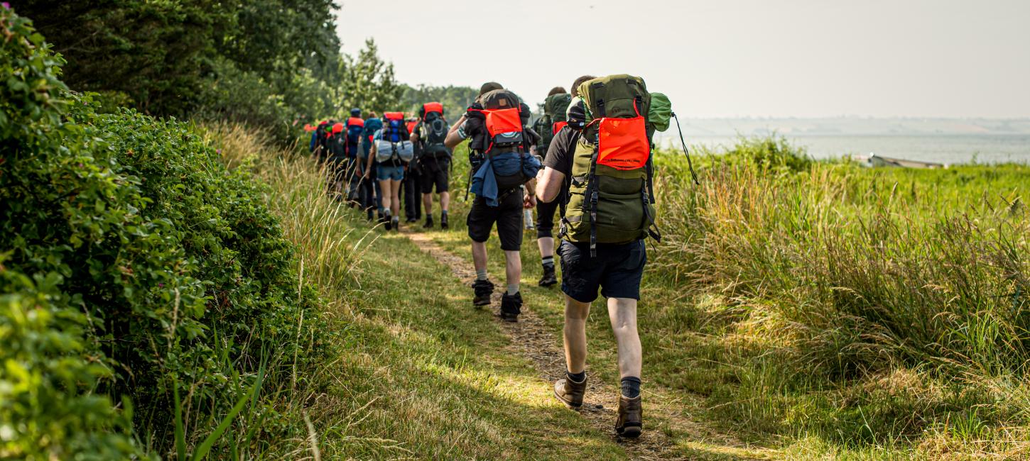 Vandrere med vandrerygsække går på en smal vandresti mellem skov og strand.