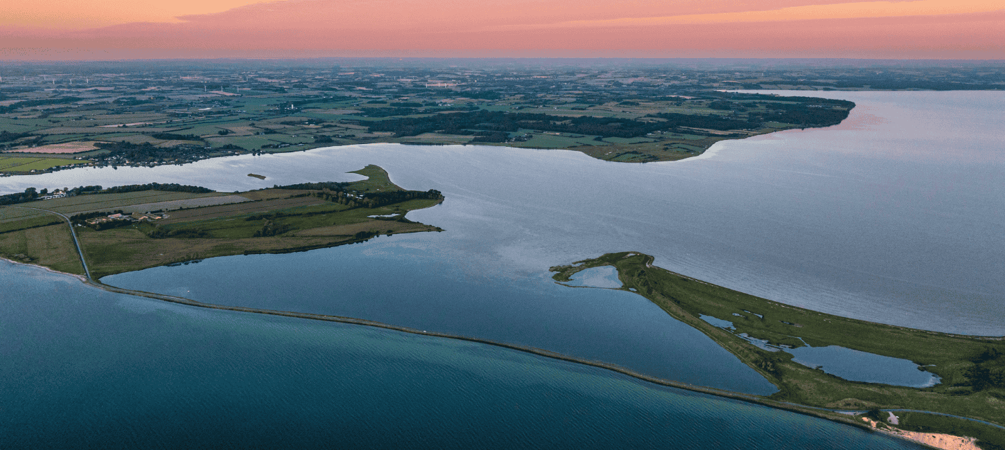 Et luttfoto af en dæmning med havvand på begge sider i solnedgang med lyserød himmel 