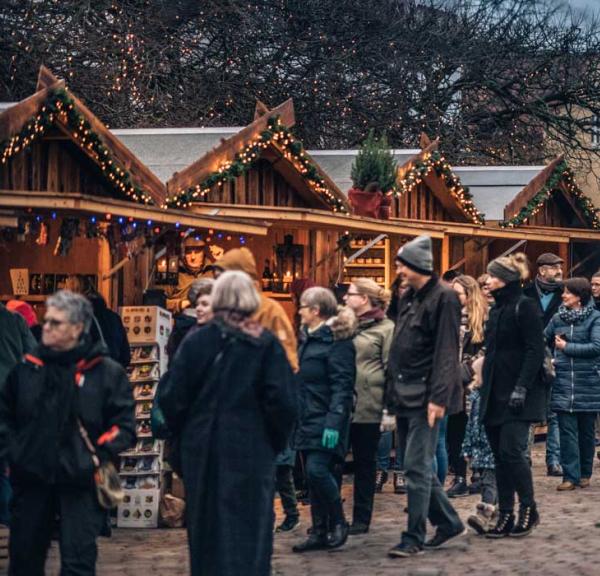En brostensplads er fyldt med mennesker pakket ind i vintertøj. Rundt om pladsen står små træhytter pyntet med gran og julelys. Træhytterne har en åben front som giver adgang til de juleting de sælger.