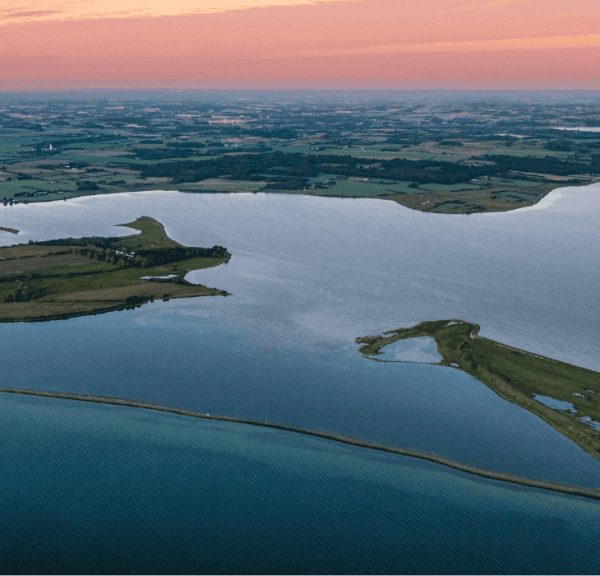 Et luttfoto af en dæmning med havvand på begge sider i solnedgang med lyserød himmel 