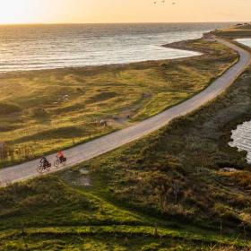 To cyklister cykler på smal grussti lige ud til vandet ved en fynsk strand. I baggrunden ses en smuk solnedgang. 