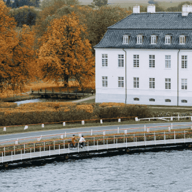 To cyklister kører på en cykelsti ude på vandet foran et hvidt gods en overskyet dag