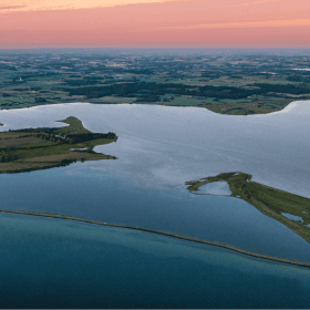 Et luttfoto af en dæmning med havvand på begge sider i solnedgang med lyserød himmel 